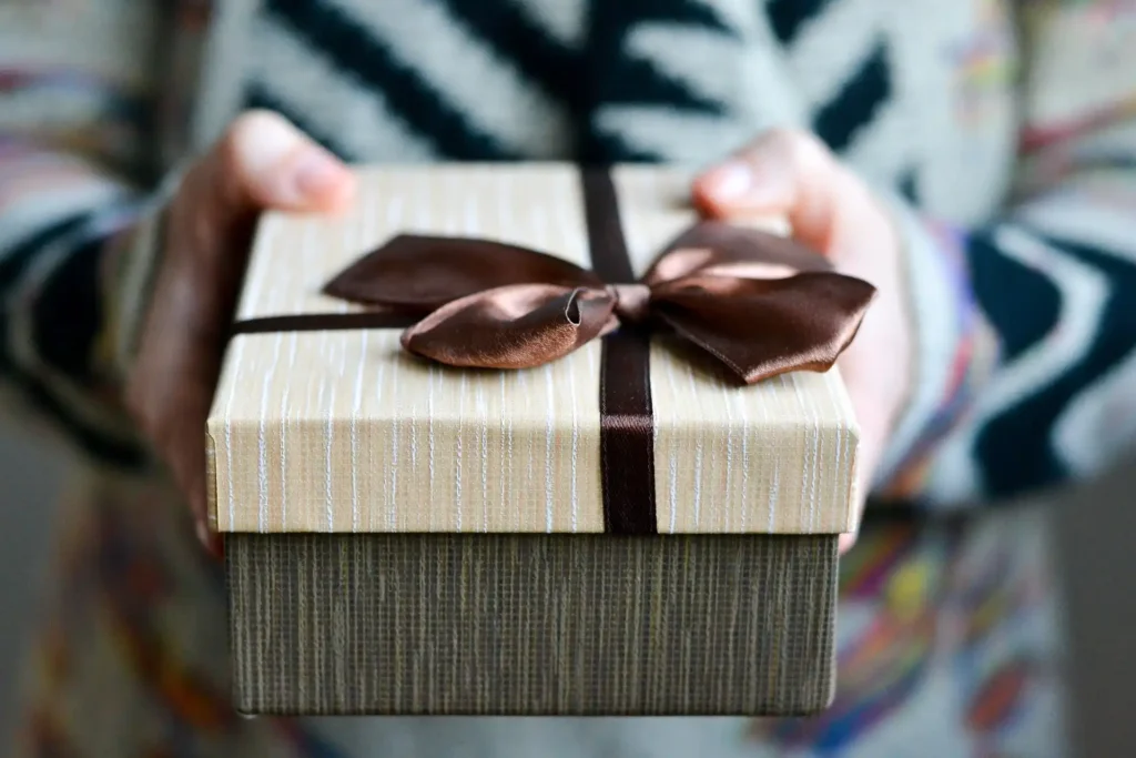 In the picture two hands are holding out a brown and beige gift package with a large brown satin bow.