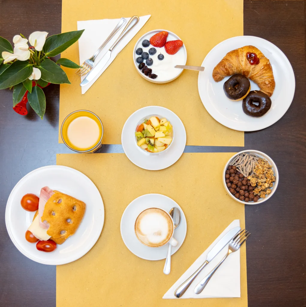In the picture, breakfast in the hotel with various products. There is a small plate with a focaccia with cooked ham, cheese and cherry tomatoes, a small plate with croissants and 2 donuts, yogurt with red fruits, fruit salad, cereals and muesli, a glass of juice and a cappuccino. A small red watering can with a plant and flowers decorates the table.